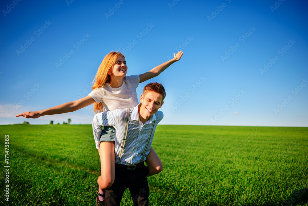 Smiling Man is holding on his back happy woman, who pulls out her arms and simulates a flight against the background of the blue sky and the green field