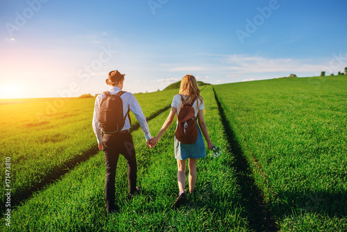 With a backpack, a man in a hat and a woman with long hair go along the path. A couple walks along the meadow