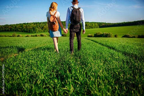 With a backpack, a man in a hat and a woman with long hair go along the path. A couple walks along the meadow