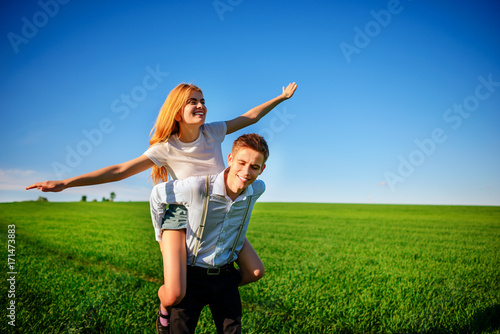 Smiling Man is holding on his back happy woman, who pulls out her arms and simulates a flight against the background of the blue sky and the green field