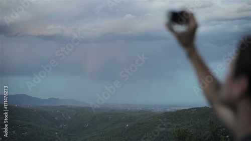 Man traveller on top of hill holds gopro camera shooting beautiful scenic twilight landscape of evening valley for blog about lifestyle travel. Dreams come true freedom inspiration life goals concept