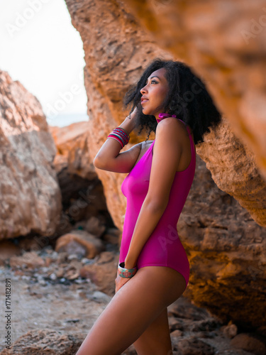 Portrait of cheerful black people, pretty happy young african american woman smiling. Rocks beach. Sexy girl in pink swimsuit and jewelry enjoying nature. Travel, holiday concept.