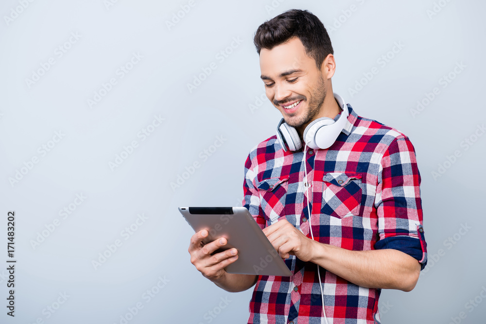 Successful young brunet freelancer hipster in checkered shirt is standing on the pure grey background with tablet, smiling, looking at it Successful young brunet freelancer hipster in checkered shirt is standing on the pure grey background with tablet, smiling, looking at it