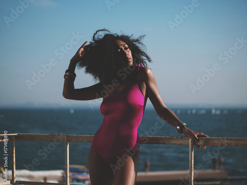Fototapeta Naklejka Na Ścianę i Meble -  Portrait of cheerful black people, pretty happy young afro american woman smiling on sea beach. Sexy girl in pink swimsuit and jewelry enjoying nature.Lady wearing bikini.
