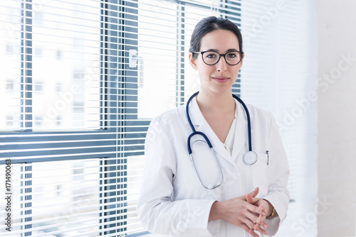 Portrait of a young nurse or physician wearing eyeglasses and white medical gown while looking at camera with confidence and serenity indoors  photo