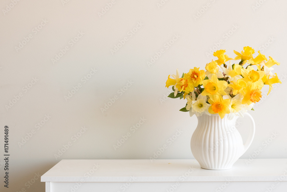 Varied yellow daffodils in white jug on table against neutral wall background