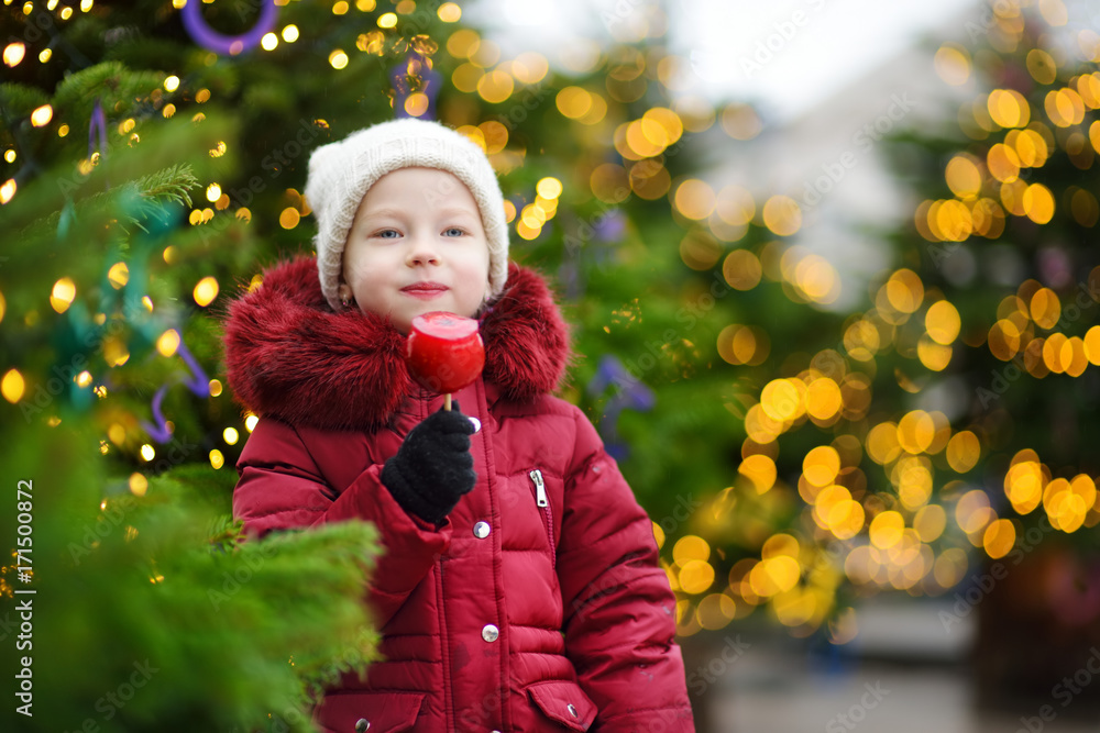 Adorable little girl eating red apple covered with sugar icing on traditional Christmas market. Adorable little girl eating red apple covered with sugar icing on traditional Christmas market.