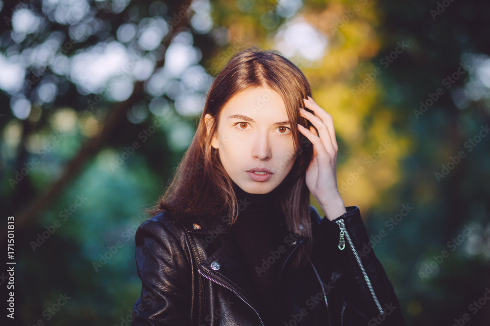 Close up art portrait of a young pretty brunette woman posing outdoors in black leather coat in golden sunlight evening spot