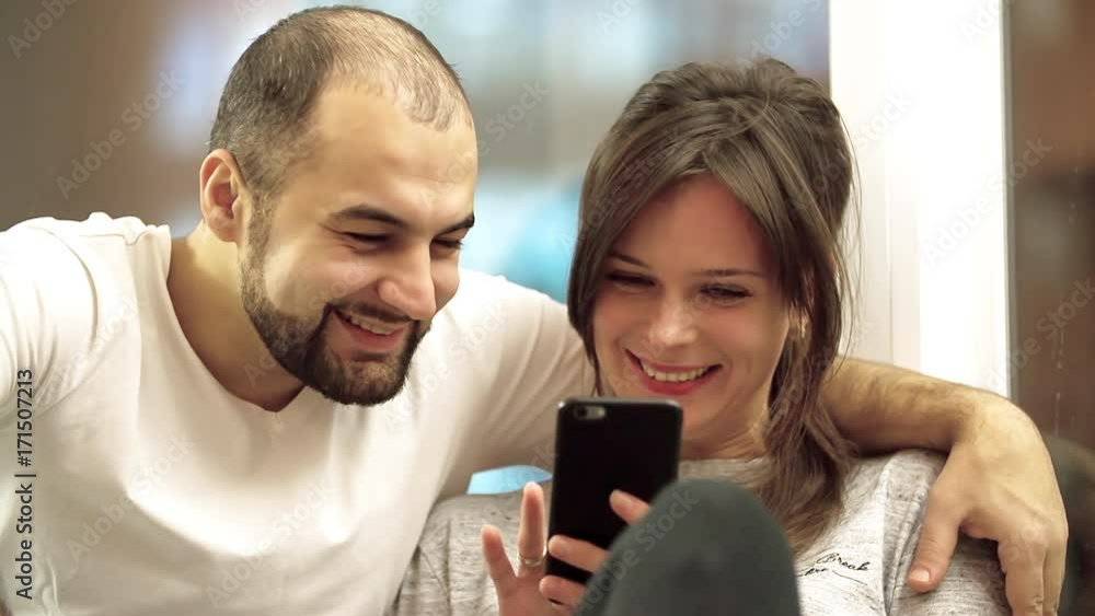 Laughing happy couple looking together at smartphone HD slow-motion video. Man and woman family smiling at home apartment in near evening window Laughing happy couple looking together at smartphone HD slow-motion video. Man and woman family smiling at home apartment in near evening window