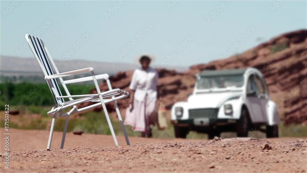 Man Unfolding Metal Beach Chairs While His Wife Walks Past Their Parked Vintage Car in the Mountains Man Unfolding Metal Beach Chairs While His Wife Walks Past Their Parked Vintage Car in the Mountains