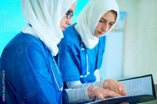 Close-up of two arabic female doctors dressed in blue uniform and white hijab sitting in a hospital reading a medical report
