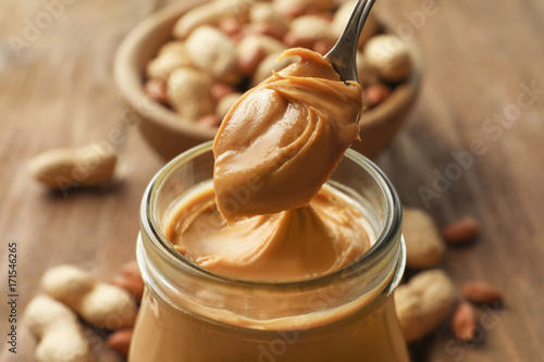 Spoon and glass jar with creamy peanut butter on kitchen table, closeup photo