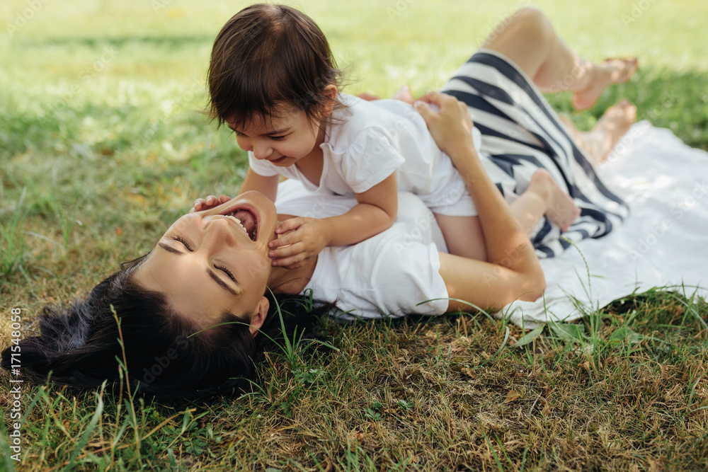 Happy Caucasian mother and her little daughter playing outdoors on grass after picnic. Cute mom and her cheerful child have fun in the park. Portrait of happy family. Motherhood and childhood concept.