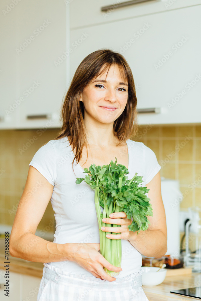 Vertical portrait brunette woman holding a green selery looking into the camera in the kitchen. Healthy eating concept. Diet.