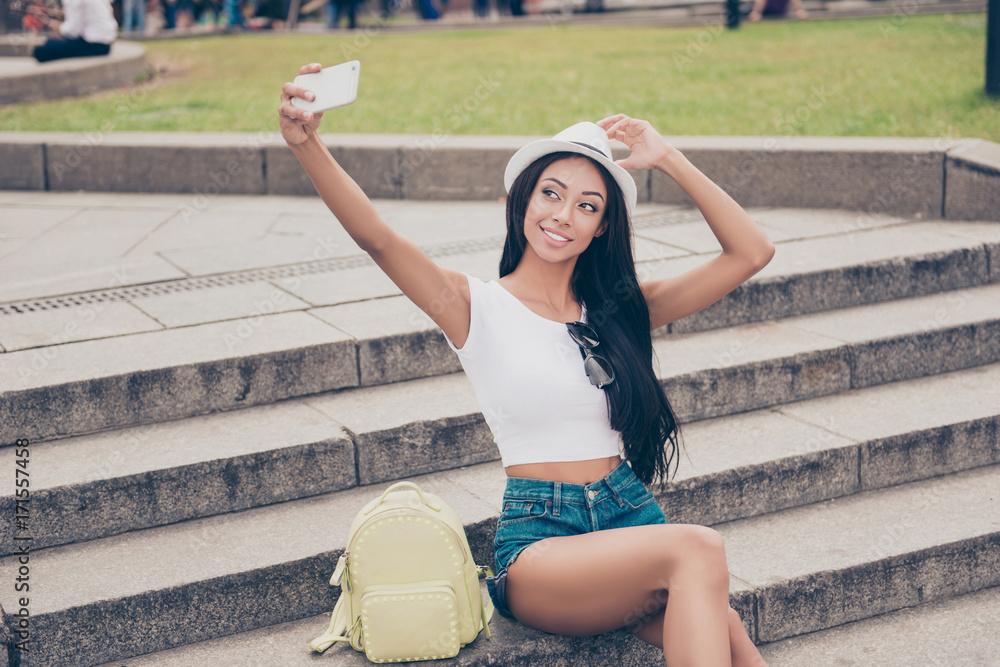 Cute lady with bronze skin and long dark hair, in cap is making a selfie shot while outside in town. She sits on concrete rung, holds headwear, in short jeans denim shorts, in white top Cute lady with bronze skin and long dark hair, in cap is making a selfie shot while outside in town. She sits on concrete rung, holds headwear, in short jeans denim shorts, in white top