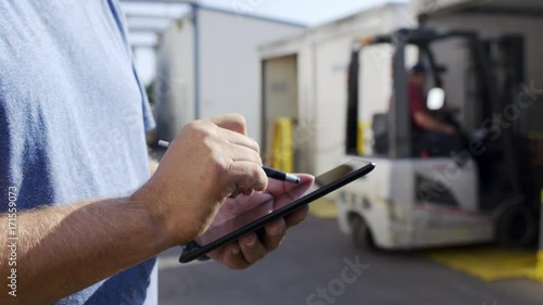 Man's hands making notes on electronic tablet while forklift driver loads a truck with pallets in background. Close up view with focus on hands, 4K