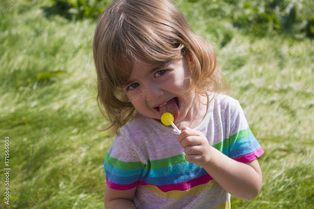 little pretty girl eating caramel lollipop on a background of green grass and smiling little pretty girl eating caramel lollipop on a background of green grass and smiling