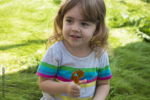 little pretty girl eating caramel lollipop on a background of green grass and smiling