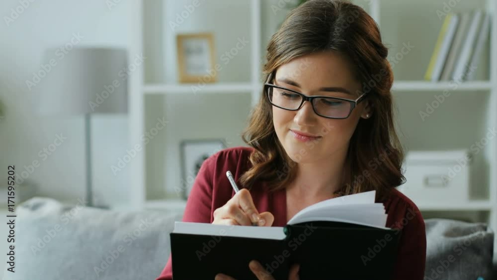 Portrait of beautiful young woman in glasses making some notes in the notebook while sitting on the sofa at home. Portrait of beautiful young woman in glasses making some notes in the notebook while sitting on the sofa at home.