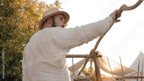 Medieval man in straw hat with beard blows the fire with the special device for wind-blowing photo