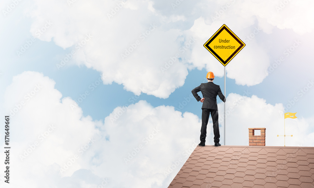 Young businessman on house brick roof holding yellow signboard. Mixed media Young businessman on house brick roof holding yellow signboard. Mixed media