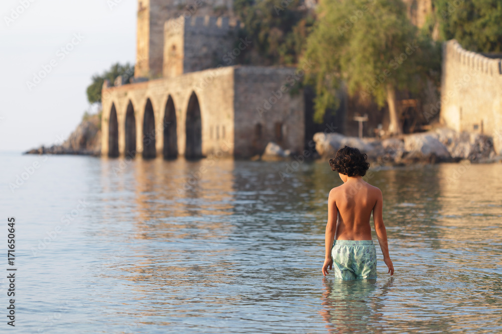 Fototapeta premium Backview portrait of a boy in swimwear near Alanya medieval ship