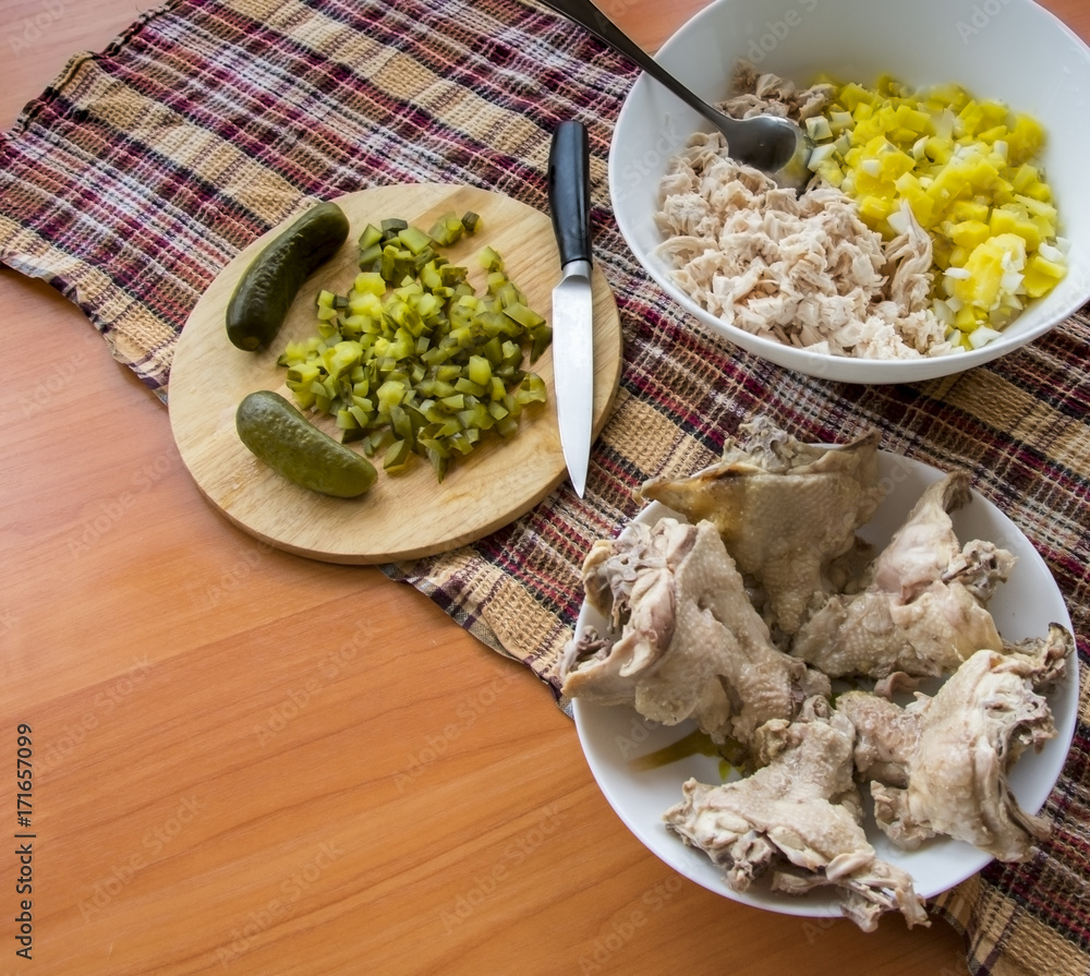 Finely chopped salad in a bowl. pickled cucumber on the board and a cutting knife; Boiled chicken backs, onions on the wooden table top. Finely chopped salad in a bowl. pickled cucumber on the board and a cutting knife; Boiled chicken backs, onions on the wooden table top.