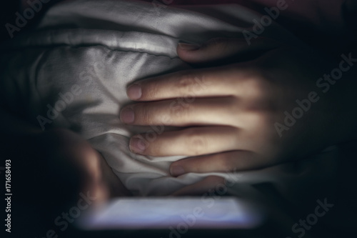 Close-up. The smartphone is held in hands. With one hand, point to the display. Lying in bed. Blurred dark background. At night
