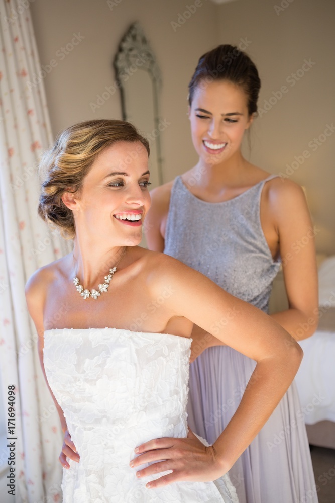 Happy bridesmaid adjusting bride wedding dress in room