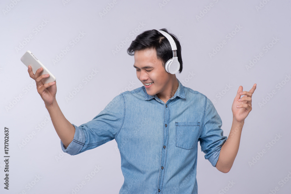 Handsome young man wearing headphones and holding mobile phone while standing against grey Handsome young man wearing headphones and holding mobile phone while standing against grey