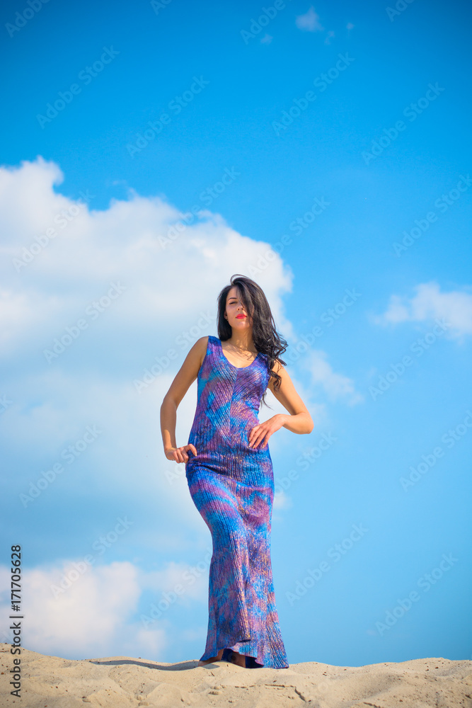 Woman with brunette curly hair in blue-purple long dress near sand dunes against sky background. Sexy stylish fashionable thin lady posing in desert Woman with brunette curly hair in blue-purple long dress near sand dunes against sky background. Sexy stylish fashionable thin lady posing in desert