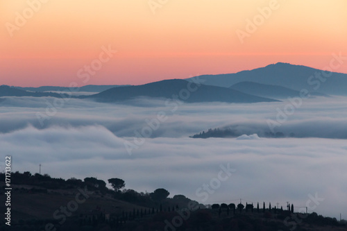 A valley filled by fog at sunset  with some hills and trees in the foreground and other hills and mountains in the foreground