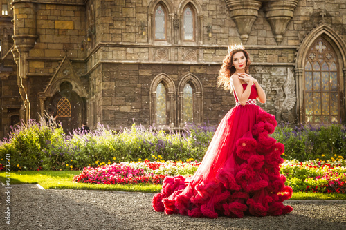 A beautiful woman, a queen in a burgundy lavish dress, walks along a flowering garden. Ancient, Gothic castle on the background. Copy space photo