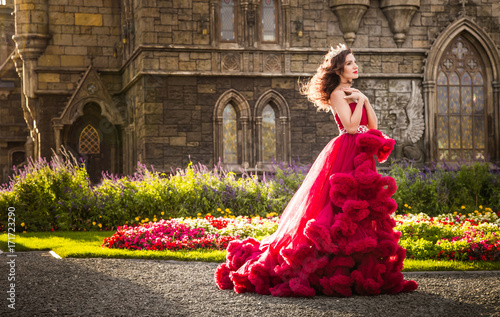 A beautiful woman, a queen in a burgundy lavish dress, walks along a flowering garden. Ancient, Gothic castle on the background. Copy space photo