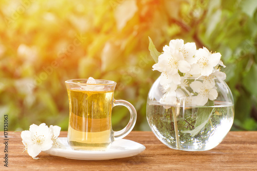 Transparent mug of tea and a vase with jasmine on a wooden table, greens on the background