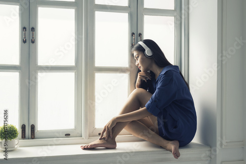 Asian women sitting listening music in wireless headphones on windowsill,Looking out the window, missing or thinking to somebody or something