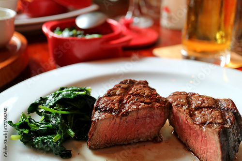 Medium grilled tenderloin steak cut in half with sauteed spinach on white plate, blurred side dishes and beverage in background 