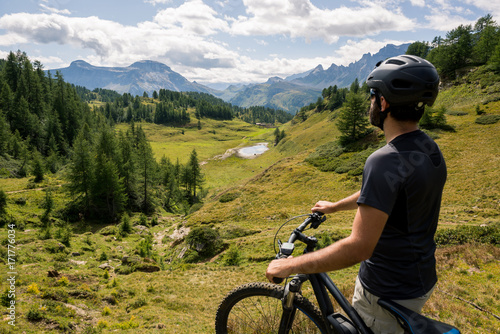 Young adult active man on mountain wearing bike helmet looking at scenic panorama holding electric bike in sunny summer day outdoor.