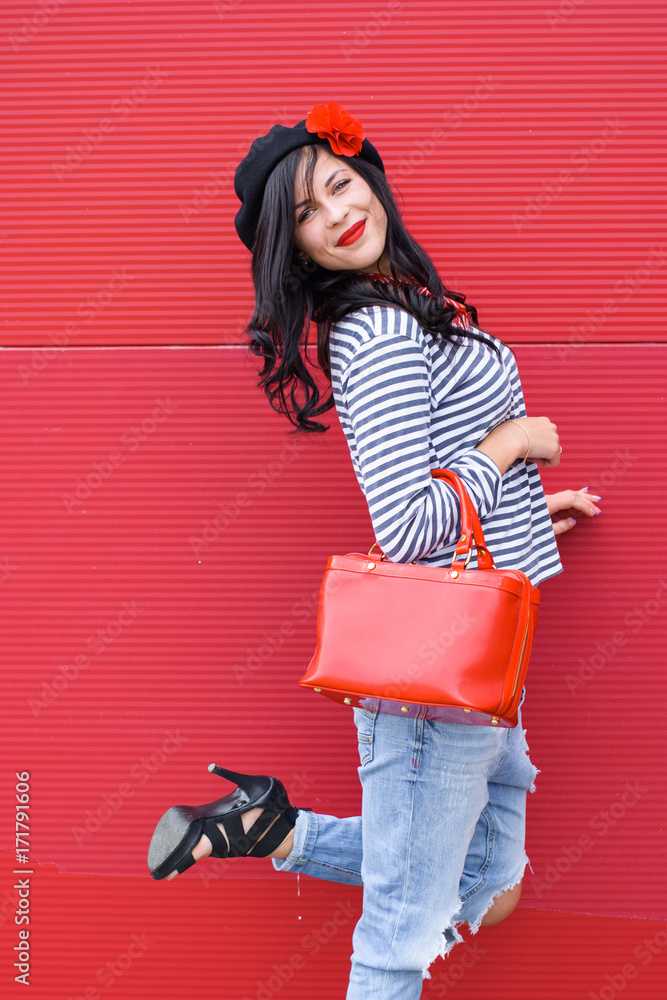 A young woman in a striped jumper, ripped jeans and beret with a flower  with a red patent leather bag. A lady in French style stands opposite the  red wall Stock Photo