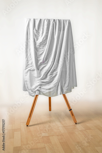 covered painting on an easel, white cloth over the picture, wooden floor and light background, art concept for an exhibition opening day or a presentation ceremony, copy space