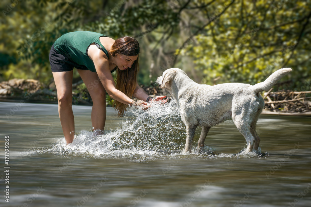 Wasserschlacht - Junge Frau spielt mit einem labrador retriever hund welpen in einem Fluss und spritzt mit Wasser