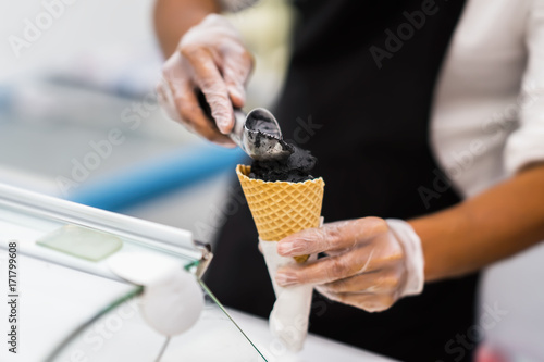 Handsome young waiter in elegant classic suit putting Black ice cream balls in waffle cone. Trendy dessert. Real scene. Professional service, catering concept