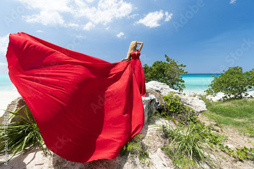Beautiful young woman with blond long hair in red fluttering dress stands on the rocks of caribbean sea