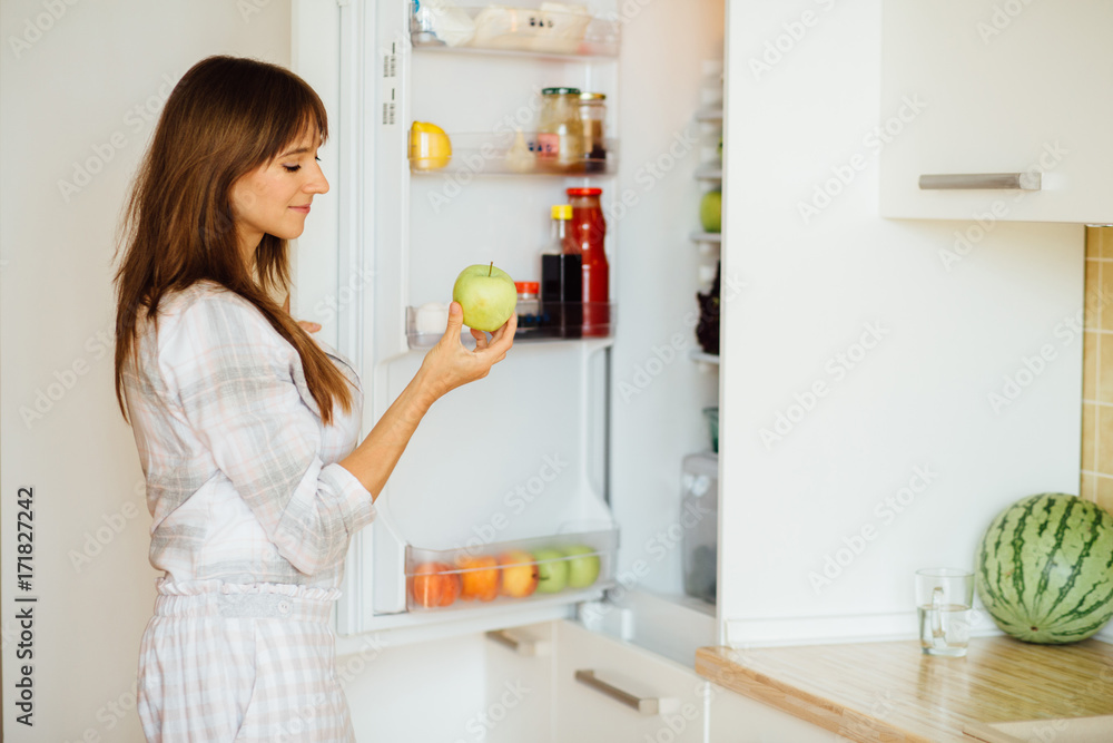 Cute female in pajamas taking apple from the fridge, attractive housewife take care about health, fresh tasty organic food, healthy eating concept.
