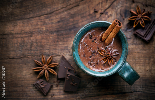 Hot chocolate in a cup with a cinnamon stick, anise star and dark chocolate flakes on rustic wooden background with an empty tag. Overhead view with copy space for your text