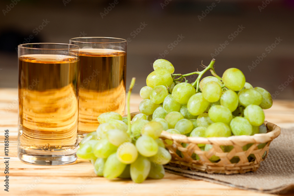 Bunches of fresh ripe green grapes in wicker basket on piece of sackcloth and two glasses with grape juice on a wooden textured backdrop. Bunches of fresh ripe green grapes in wicker basket on piece of sackcloth and two glasses with grape juice on a wooden textured backdrop.
