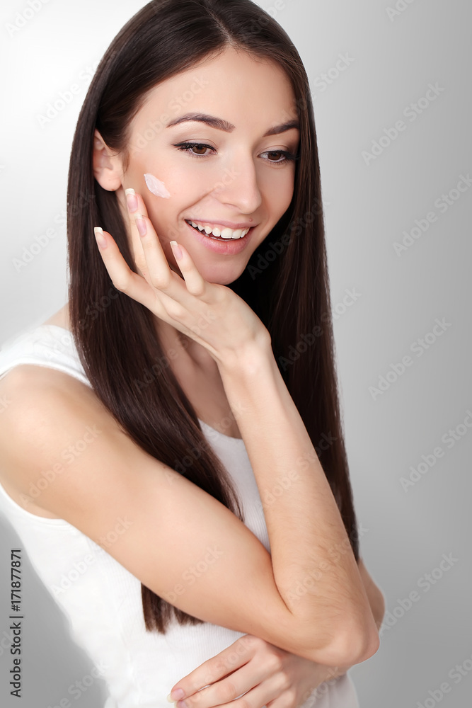 Beautiful young woman applying face cream to her cheek bone in a beauty skincare or cosmetics concept on grey with copy space Beautiful young woman applying face cream to her cheek bone in a beauty skincare or cosmetics concept on grey with copy space