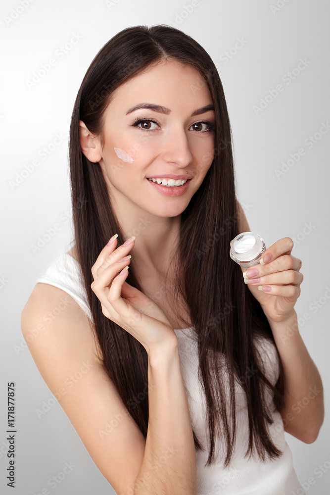 Beautiful young woman applying face cream to her cheek bone in a beauty skincare or cosmetics concept on grey with copy space Beautiful young woman applying face cream to her cheek bone in a beauty skincare or cosmetics concept on grey with copy space