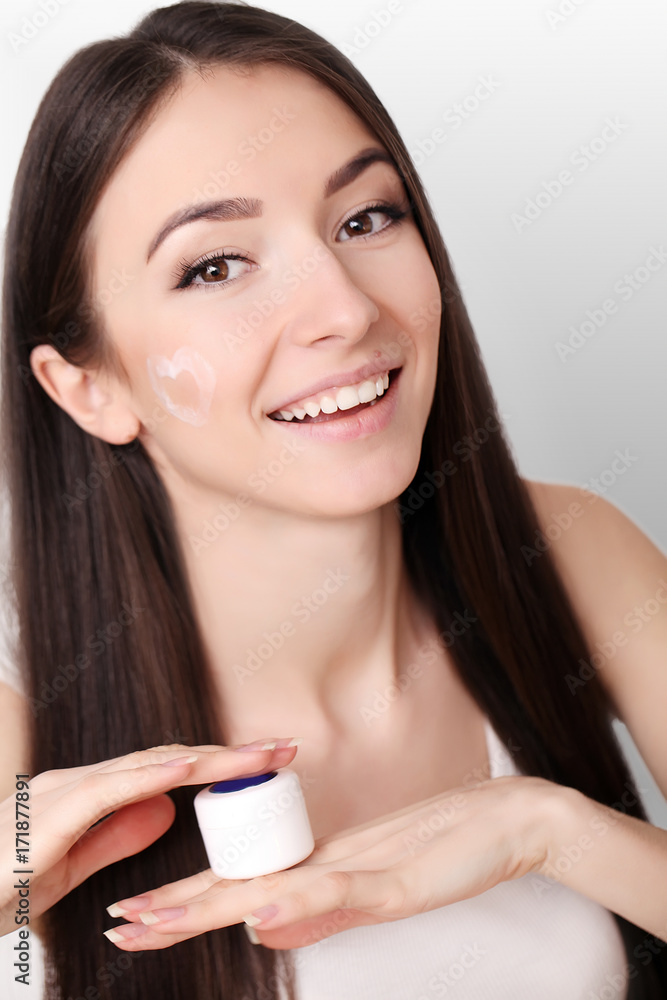 Beautiful young woman applying face cream to her cheek bone in a beauty skincare or cosmetics concept on grey with copy space Beautiful young woman applying face cream to her cheek bone in a beauty skincare or cosmetics concept on grey with copy space