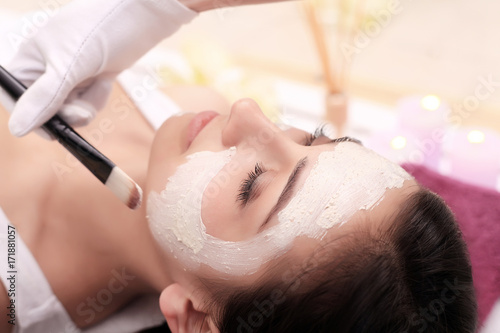 Beautiful young woman is getting facial clay mask at spa, lying with cucumbers on eyes